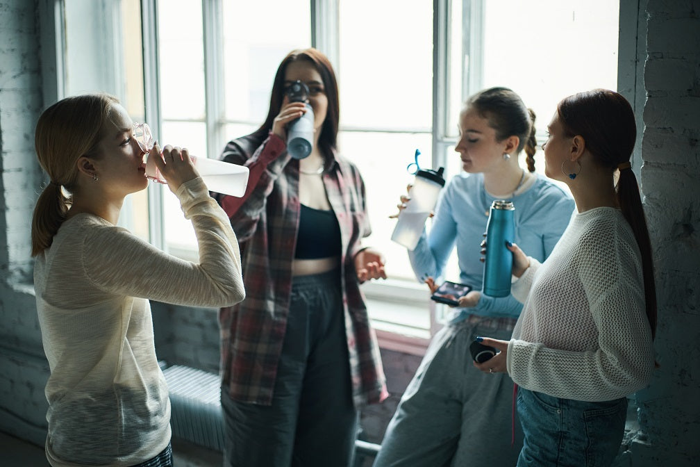 Group of young women drinking water while standing near large windows, engaging and relaxed. Hydrating and sharing a moment during their routine