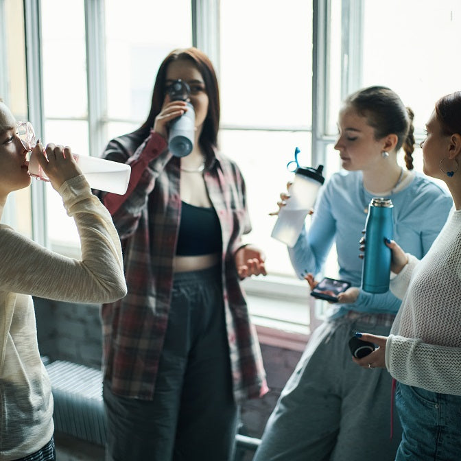 Group of young women drinking water while standing near large windows, engaging and relaxed. Hydrating and sharing a moment during their routine